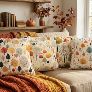 May include: Four square throw pillows arranged on a sofa, each featuring a cream-colored fabric printed with a repeating pattern of stylized trees in shades of orange, yellow, brown, and grey. A textured orange throw blanket is draped over the sofa.
