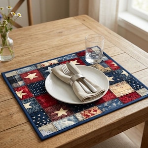 May include: A rectangular placemat with a patchwork design in red, white, and blue, featuring stars and stripes. A white plate with a folded beige napkin, fork, and knife, and a clear glass are on the placemat. The placemat is on a wooden table.