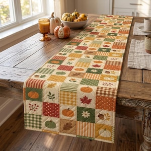 Puede incluir: Un camino de mesa acolchado con un diseño de patchwork de cuadrados con patrones de temática otoñal que incluyen calabazas, hojas de arce, bellotas y estampados de cuadros en tonos naranja, verde, amarillo y marrón sobre una mesa.
