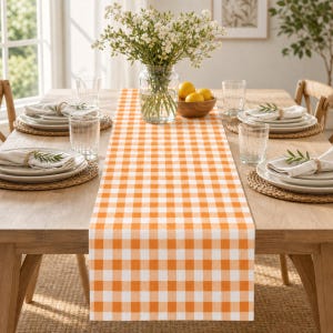 May include: A rectangular table runner featuring a vibrant orange and white gingham checkered pattern. The runner is laid flat across the center of a wooden dining table, which is set with white plates, clear glasses, and woven placemats.