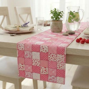 May include: A pink patchwork table runner with a variety of patterns, including polka dots and floral prints, is displayed on a light-colored wooden table. The runner is decorated with squares of varying shades of pink and white. A pitcher of water and a small potted plant are also on the table.