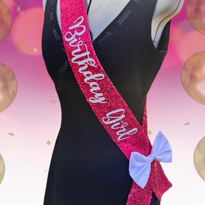 May include: A hot pink sequined sash with the words "Birthday Girl" in white script. The sash is adorned with a white bow and is draped over a black dress form. The background features pink and gold balloons.