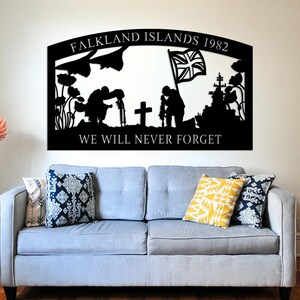 Puede incluir: Una pieza de arte mural de metal negro con el texto "FALKLAND ISLANDS 1982" y "WE WILL NEVER FORGET". La silueta representa soldados, una cruz y un barco. El arte cuelga sobre un sofá gris con cojines estampados.