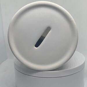 May include: White ceramic money box with a slot on the top. The money box is sitting on a white base.