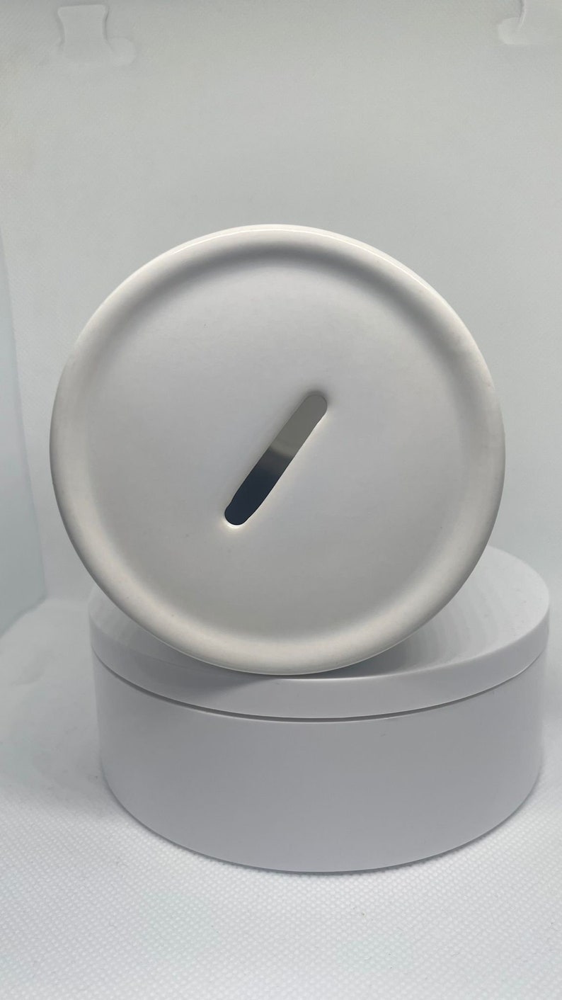 May include: White ceramic money box with a slot on the top. The money box is sitting on a white base.
