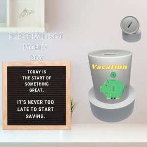 May include: A white ceramic money box with a slot on top. The money box has a green piggy design with dollar signs on it. The money box is labelled "Vacation" in yellow letters. The money box is sitting on a white base. A motivational message is displayed on a letter board behind the money box.