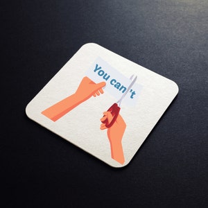 K&ouml;nnte beinhalten: Ein wei&szlig;er Untersetzer mit einer Cartoon-Illustration einer Hand, die eine Schere h&auml;lt und das Wort "You can't" von einem St&uuml;ck Papier schneidet.