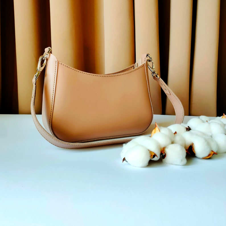 May include: A light brown leather shoulder bag with a curved shape and a silver clasp. The bag is sitting on a white surface with white cotton balls in the background.