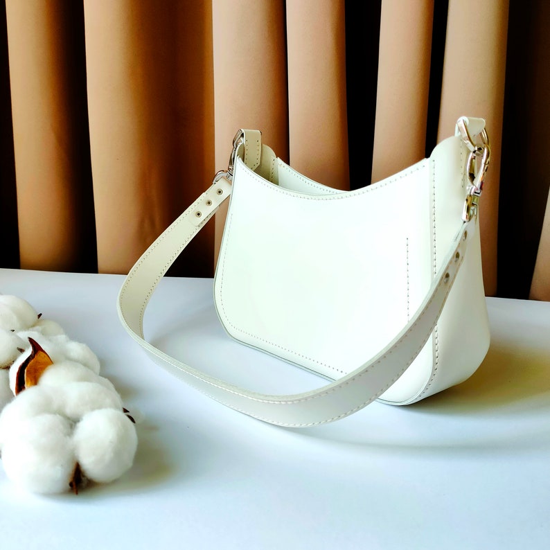 May include: A white leather shoulder bag with a curved shape and a single strap. The bag is on a white surface.