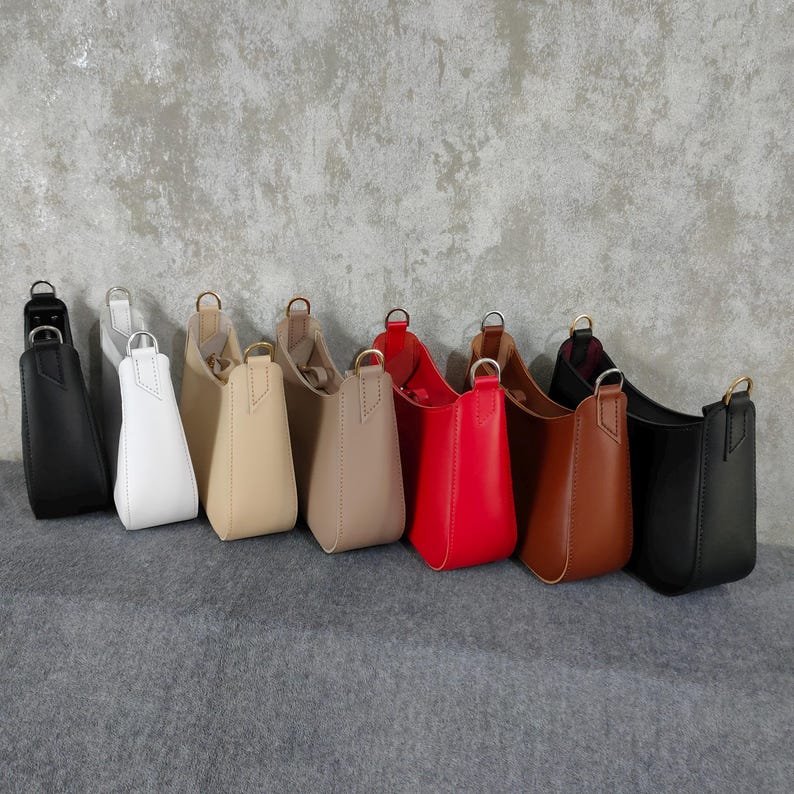 May include: Eight leather handbags in different colors, including black, white, beige, brown, and red. The handbags are arranged in a row on a gray textured surface.