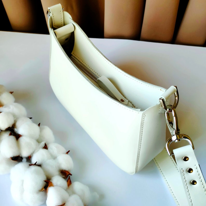 May include: A white leather shoulder bag with a zipper closure and a long adjustable strap. The bag is made of a soft, supple leather and has a classic, elegant design.