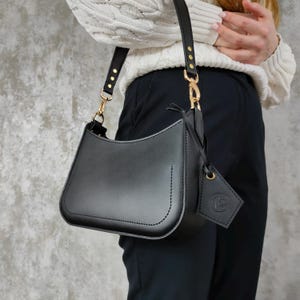 May include: A smooth black leather shoulder bag with a curved silhouette, featuring gold-tone metal hardware, a single adjustable strap with gold rivets, and a matching black leather luggage tag attached to the side with a gold-tone metal clasp.