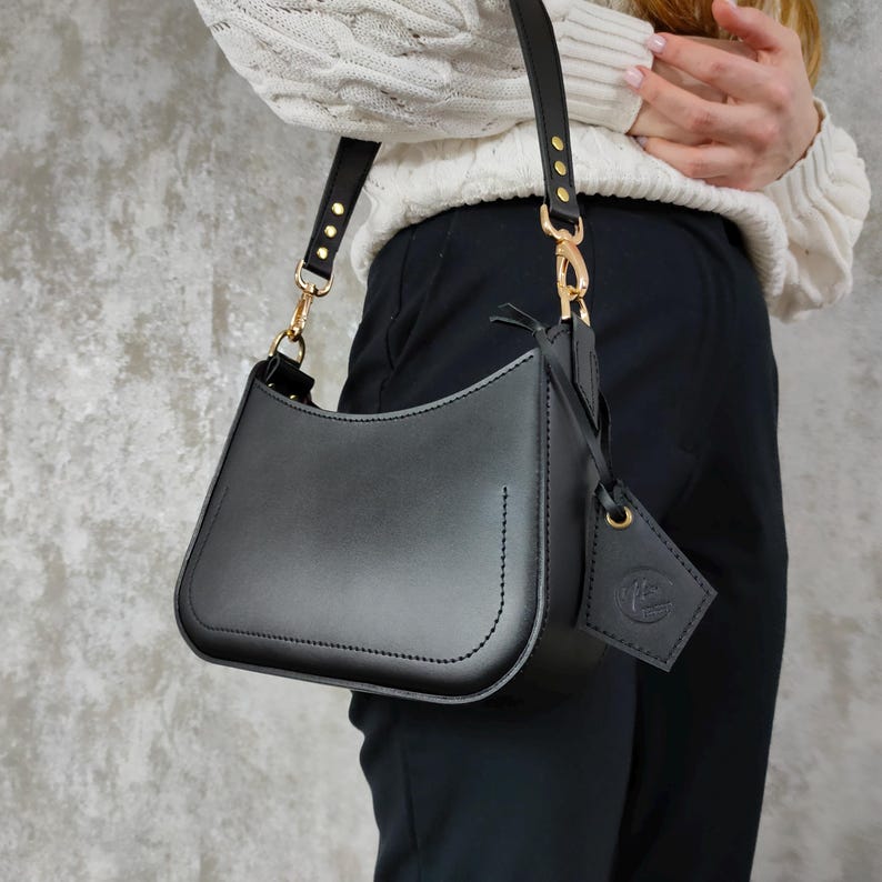 May include: A smooth black leather shoulder bag with a curved silhouette, featuring gold-tone metal hardware, a single adjustable strap with gold rivets, and a matching black leather luggage tag attached to the side with a gold-tone metal clasp.