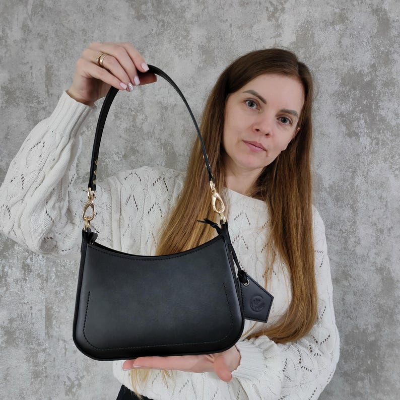May include: A black shoulder bag with a smooth finish, featuring a curved top edge, a single strap attached with gold-toned metal clasps, and a small matching black leather-like tag hanging from the side of the bag near the hardware.