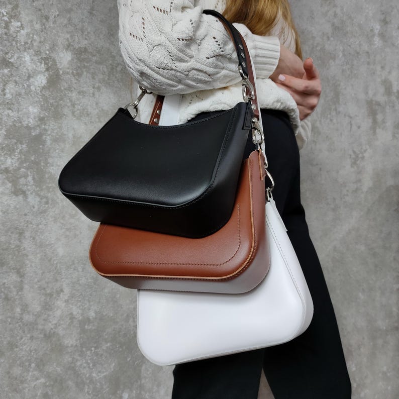 May include: Three leather handbags in black, brown, and white. The bags are stacked on top of each other, with the black bag on top. The bags have a curved shape and a single strap.