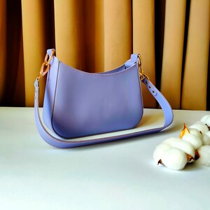 May include: A lavender-colored leather handbag with a gold-toned buckle and a long strap. The bag is sitting on a white surface.