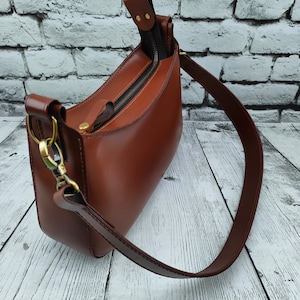 May include: A smooth brown leather shoulder bag featuring a top zipper closure with a matching leather pull tab, antique brass-toned metal hardware, and a wide adjustable strap attached with swivel lobster clasps on both sides of the bag.