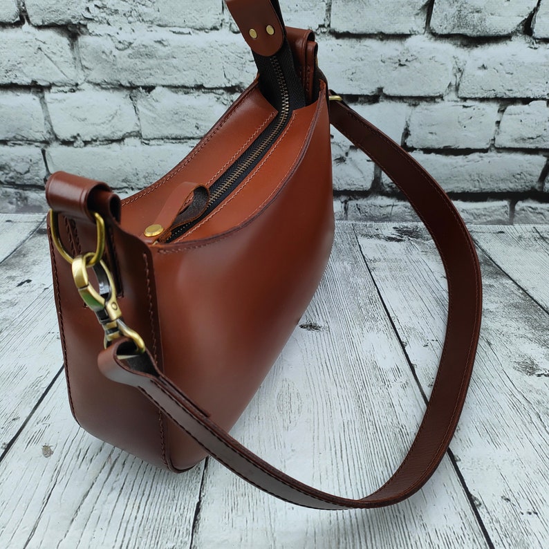 May include: A smooth brown leather shoulder bag featuring a top zipper closure with a matching leather pull tab, antique brass-toned metal hardware, and a wide adjustable strap attached with swivel lobster clasps on both sides of the bag.