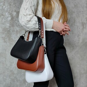 May include: Three leather handbags in black, brown, and white. The bags are all the same style, with a curved top and a single strap. The bags are hanging from a single strap.