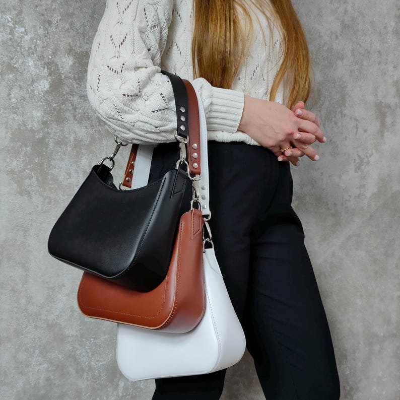 May include: Three leather handbags in black, brown, and white. The bags are all the same style, with a curved top and a single strap. The bags are hanging from a single strap.