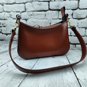 May include: A smooth, medium-brown leather shoulder bag with a curved top silhouette. The bag features a top zipper closure, a matching leather strap, and antique brass-toned metal hardware clips that attach the strap to the sides of the bag.