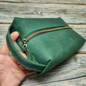 Könnte beinhalten: Eine grüne Leder-Reisetasche mit einem Lederriemen als Griff. Die Tasche hat einen goldenen Reißverschluss und der Riemen ist mit dem Wort "SMERK" geprägt.