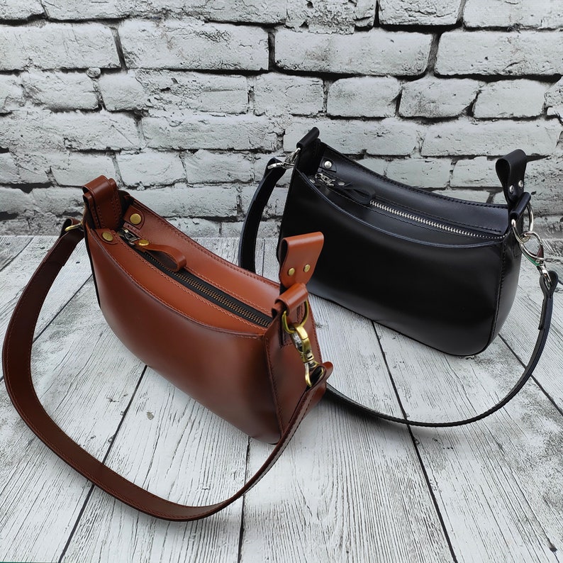 May include: Two structured shoulder bags, one in a rich brown and one in a deep black, each featuring a smooth finish, a top zipper closure with a matching pull tab, and a wide adjustable strap attached with metallic hardware on both sides.