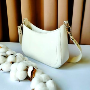 May include: A white leather handbag with a curved shape and a long strap. The bag is sitting on a white surface with cotton balls.