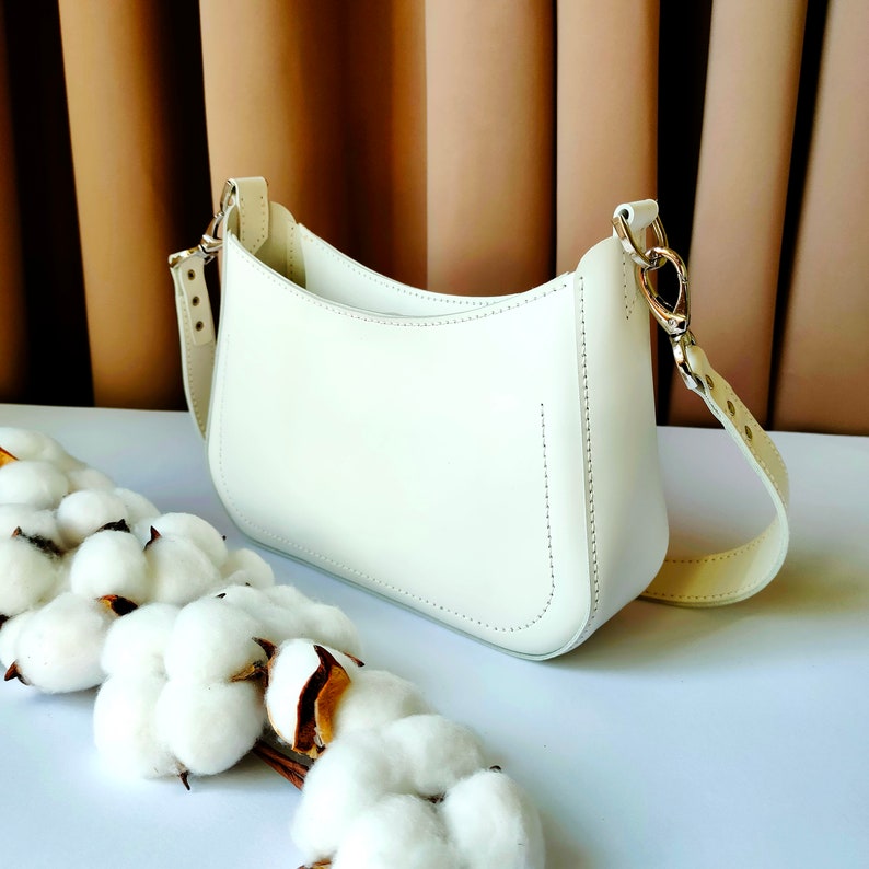 May include: A white leather handbag with a curved shape and a long strap. The bag is sitting on a white surface with cotton balls.