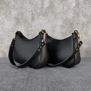 May include: Two black shoulder bags with a smooth finish, featuring curved silhouettes and wide straps attached with silver-toned metal hardware. Each bag has a structured base and a minimalist design with visible stitching along the strap attachments.