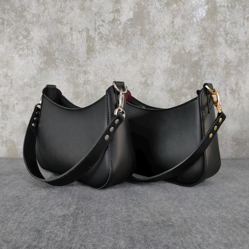 May include: Two black shoulder bags with a smooth finish, featuring curved silhouettes and wide straps attached with silver-toned metal hardware. Each bag has a structured base and a minimalist design with visible stitching along the strap attachments.