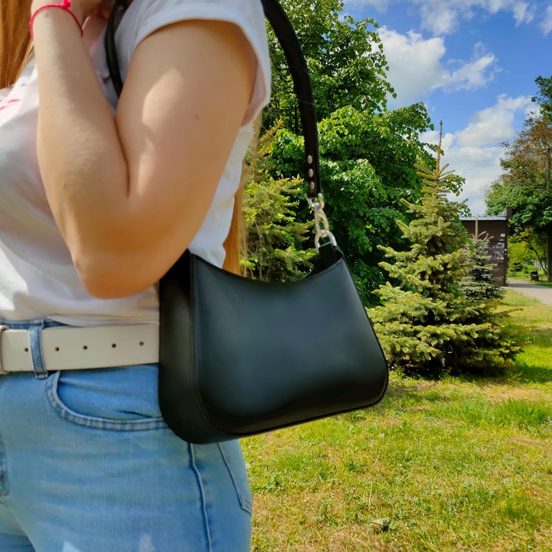 May include: A smooth black shoulder bag with a curved silhouette and a single strap attached by silver-toned metal hardware. The bag features a clean, minimalist design with a structured base and a top zipper closure, worn over the shoulder.