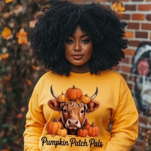 Puede incluir: Sudadera amarilla mostaza con un diseño de vaca y calabazas. El texto "Pumpkin Patch Pals" está impreso en la parte inferior. La persona que lleva la sudadera tiene el pelo rizado oscuro. El fondo incluye hojas de otoño.