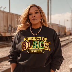Può includere: Felpa nera con la scritta "PRODUCT OF BLACK HISTORY" in un design a scacchi giallo, rosso, verde e nero. La felpa ha un girocollo e maniche lunghe. Sono visibili anche una collana a catena dorata e orecchini a cerchio.