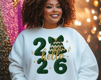 May include: A white sweatshirt with the text "26 hello 26" in green and gold. The design includes a bell and stars. The person in the image is smiling and has curly brown hair.