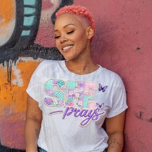 Könnte beinhalten: Weißes T-Shirt mit dem Text "SHE prays" in einem Patchwork-Design. Die Buchstaben sind mit Blumen- und Vichy-Mustern verziert, und das Wort "prays" ist in einer Schreibschrift gehalten. Zwei lila Schmetterlinge sind ebenfalls zu sehen.