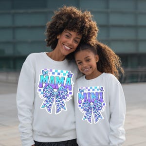 May include: Matching white sweatshirts with the words "MAMA" and "MINI" in a blue and purple gradient, with a bow design. The sweatshirts have a checkered pattern and butterfly accents.
