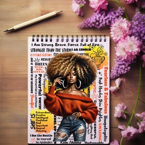 Puede incluir: Un cuaderno de espiral con una ilustración de mujer, rodeado de flores rosas y moradas. El cuaderno tiene el texto "I Am Strong. Brave. Fierce. Full of Fire." Un lápiz descansa sobre la superficie de madera.