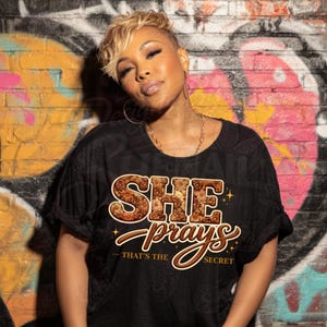Könnte beinhalten: Schwarzes T-Shirt mit dem Schriftzug "SHE PRAY'S - THAT'S THE SECRET" in Braun und Gold. Die Person trägt das T-Shirt, hat kurze blonde Haare und Goldschmuck. Der Hintergrund ist eine Backsteinmauer mit Graffiti.