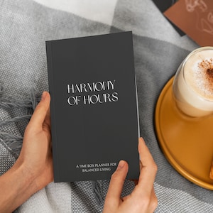 May include: A dark gray planner titled "HARMONY OF HOURS" is held open by two hands. The text "A TIME BOX PLANNER FOR BALANCED LIVING" is below the title. A glass of latte and a croissant sit on a yellow plate.