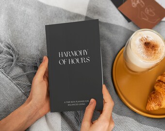 May include: A dark gray planner titled "HARMONY OF HOURS" is held open by two hands. The text "A TIME BOX PLANNER FOR BALANCED LIVING" is below the title. A glass of latte and a croissant sit on a yellow plate.