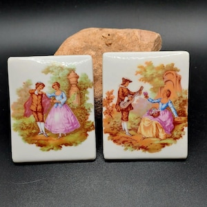 以下が含まれることがあります： 庭園の風景の中で、手描きで描かれたカップルのシーンが描かれた、ヴィンテージの磁器タイル2枚。最初のタイルには踊っているカップルが描かれ、2枚目のタイルにはギターを持ったカップルが描かれている。