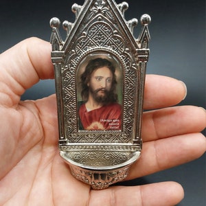 May include: A silver metal religious shrine with a small image of Jesus inside. The image has the text "¡Amigo que nunca falla!" below it.
