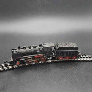 Puede incluir: Un modelo de tren negro con ruedas y detalles rojos, que incluye una locomotora y un vagón de carbón, se encuentra sobre una vía en miniatura. El tren es una réplica detallada de una locomotora de vapor clásica.