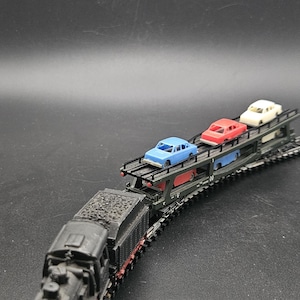 Puede incluir: Un modelo de tren negro con un vagón de carbón y un portacoches cargado con coches de juguete azules, rojos y blancos. El tren está sobre una superficie negra.
