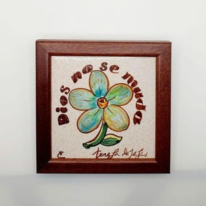 May include: A framed ceramic tile featuring a hand-painted blue flower and the text "Dios no se muda" in brown lettering. The tile is signed "Teresa de Jesús" in the bottom right corner.