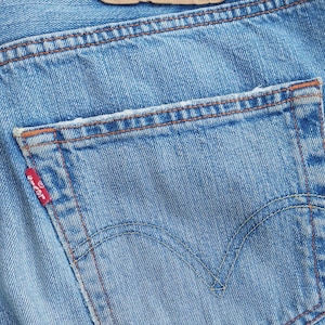 Puede incluir: Un par de pantalones vaqueros de mezclilla azul claro con un parche de cuero marrón de Levi's que dice "Levi Strauss & Co. San Francisco Cal Original  Riveted Quality Clothing XX Trade Mark Patented May 20 1873 501 W32 L32".