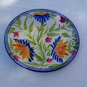 May include: A white ceramic plate with a blue rim and a floral design. The flowers are painted in shades of yellow, orange, pink, and blue.