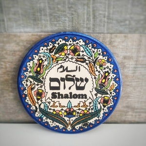 Peut inclure: Une assiette ronde en céramique avec un fond bleu et un centre blanc. L'assiette est décorée de motifs floraux colorés et des mots "Shalom" en hébreu et en anglais.