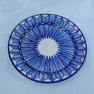 May include: A round ceramic plate with a white center and a blue geometric design. The plate's rim features a repeating pattern of blue leaf-like shapes, and a blue border. The plate is on a light blue surface.